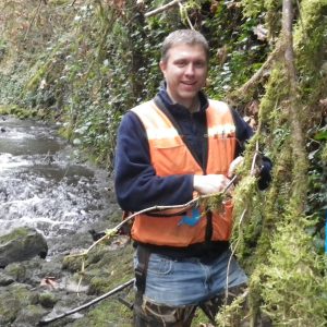 Dusty Day standing near a stream wearing an orange work vest and waders.