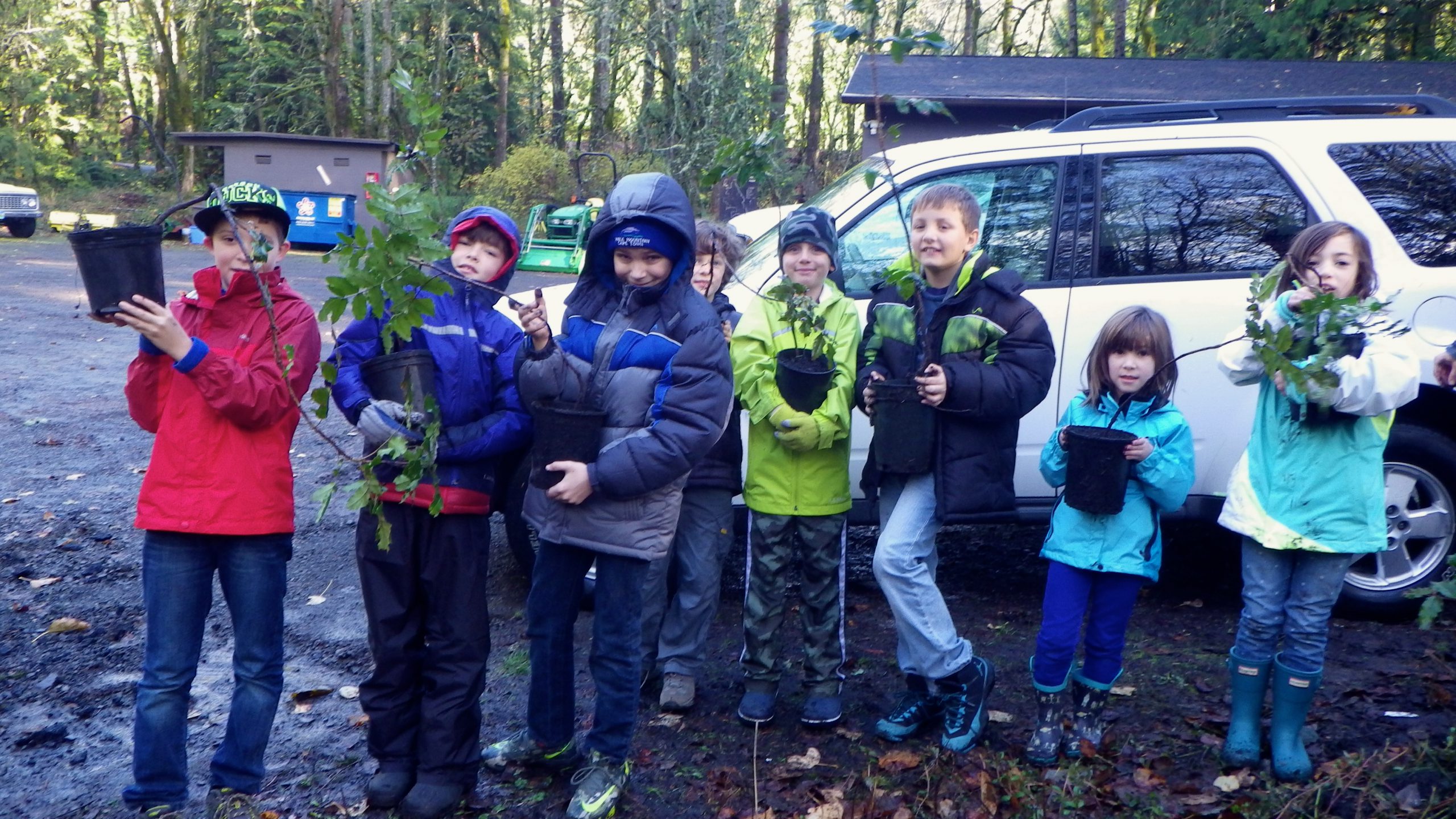 Group of 8 children outdoors wearing jackets holding potted native plants.